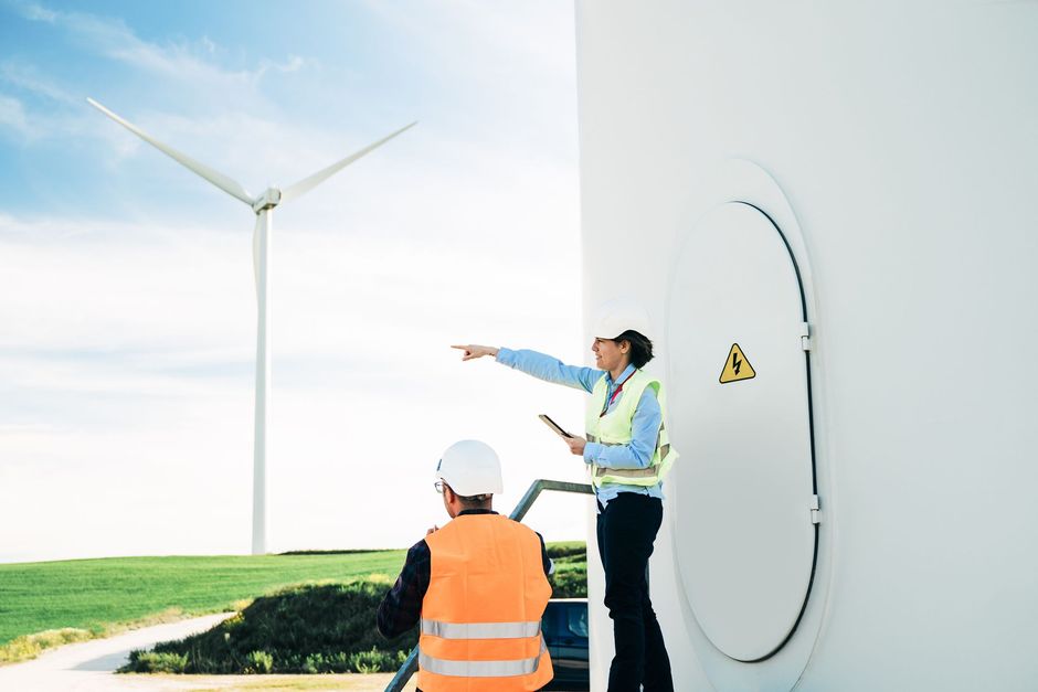 Deux ingénieurs près d'une éolienne : l'un montre du doigt, l'autre regarde ; éolienne blanche et ciel bleu en arrière-plan.