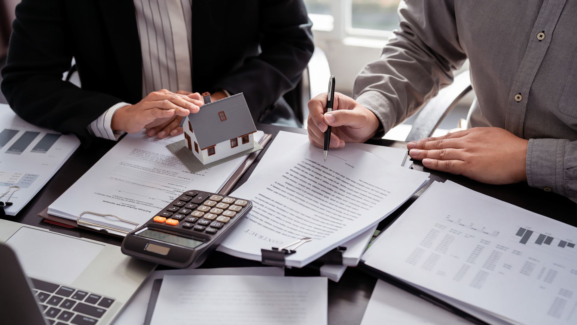 Deux personnes assises à une table examinent des documents ; une calculatrice et une petite maquette de maison sont présentes.
