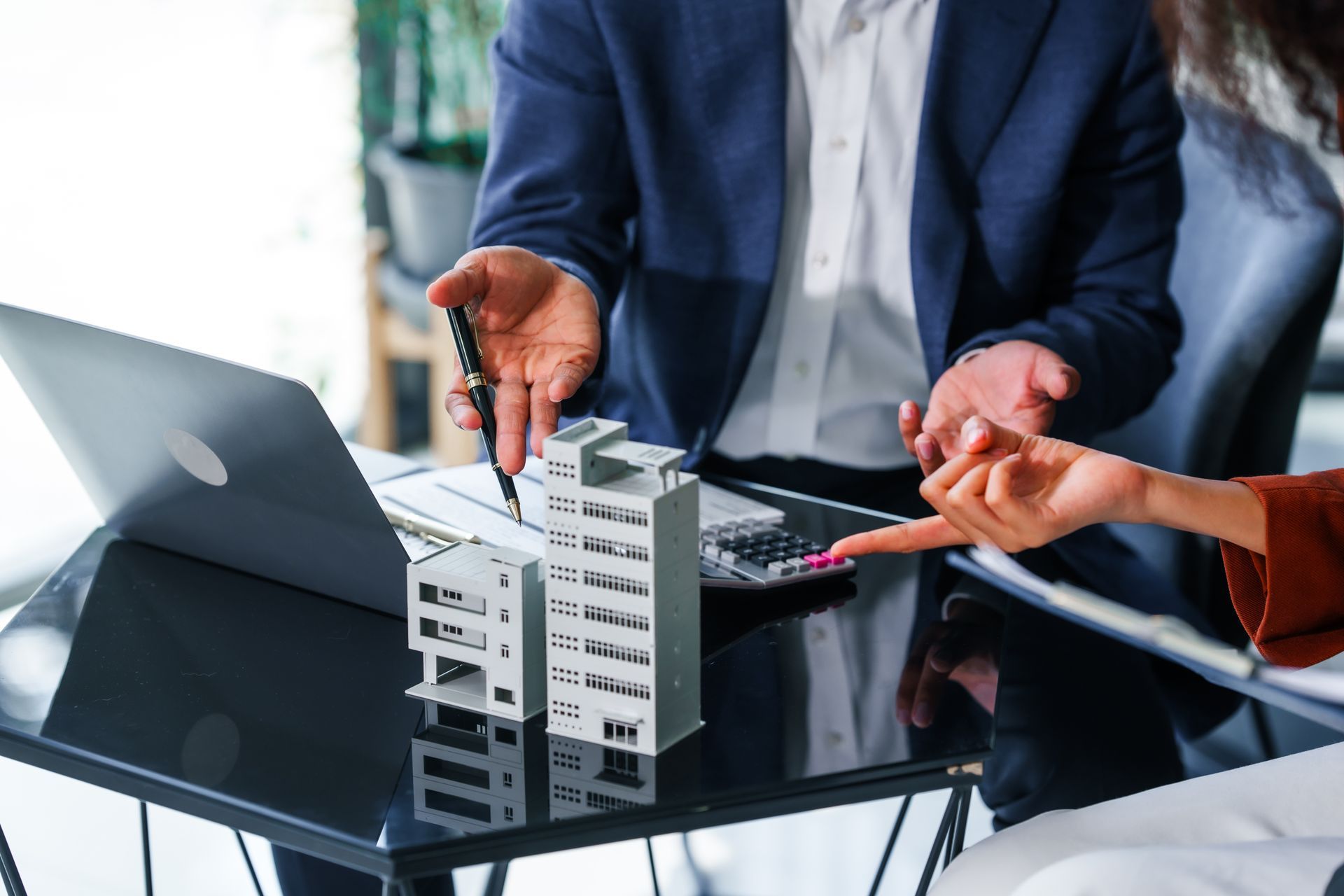 Deux personnes désignent du doigt une maquette de bâtiment miniature posée sur une table avec un ordinateur portable et une calculatrice.