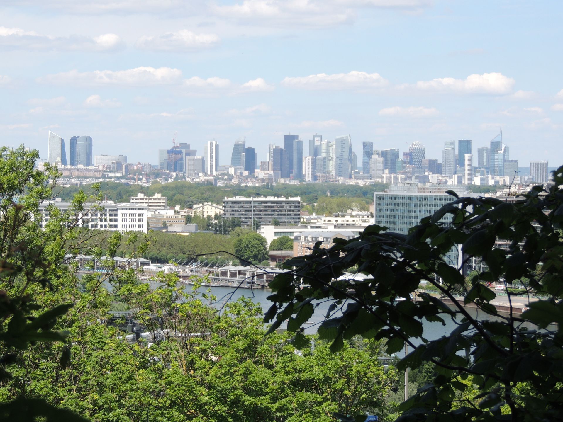 Vue sur la silhouette d'une ville avec de hauts immeubles, certains partiellement masqués par les arbres. Ciel bleu nuageux.