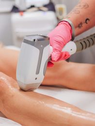 Perna de uma pessoa sendo tratada com um aparelho de depilação a laser. A mão com luva rosa opera o aparelho.