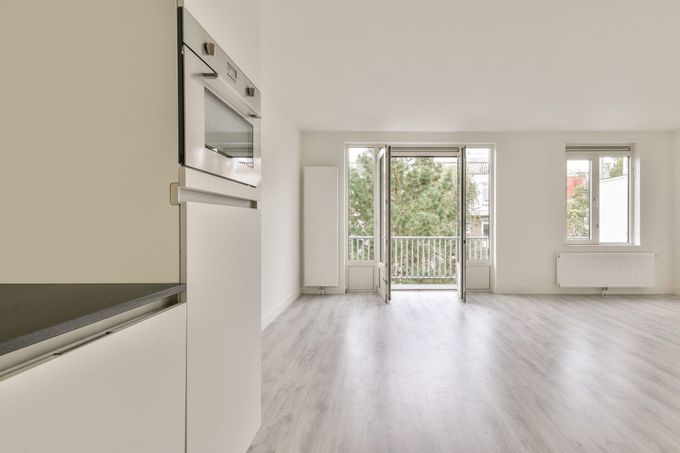 Sala de estar luminosa y vacía en un apartamento, con suelos de madera clara, paredes blancas, puertas que dan al balcón y una ventana.