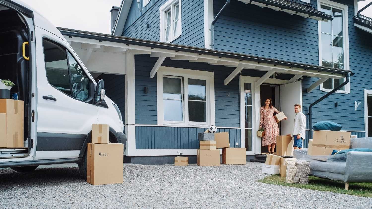 Dos personas desempaquetando cajas afuera de una casa azul junto a una furgoneta blanca.