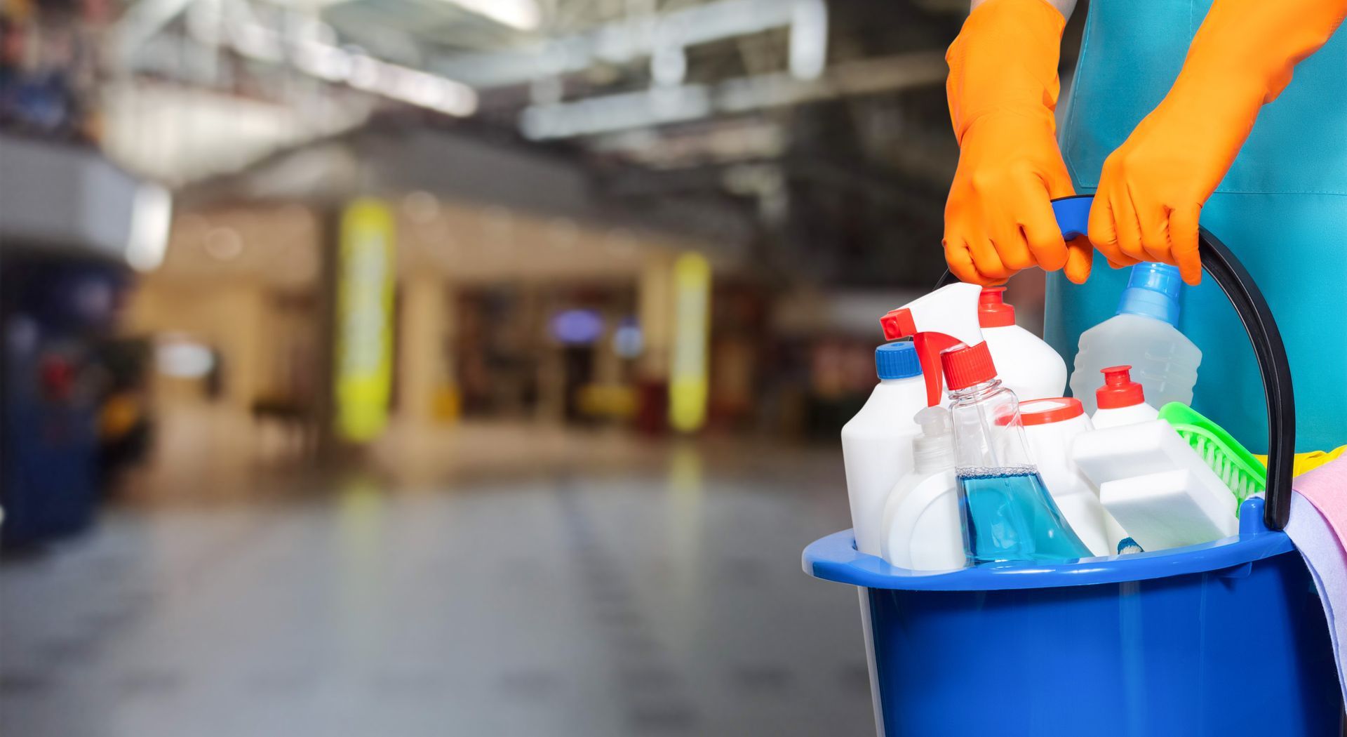 Una persona que lleva guantes naranjas sostiene un balde azul lleno de productos de limpieza.