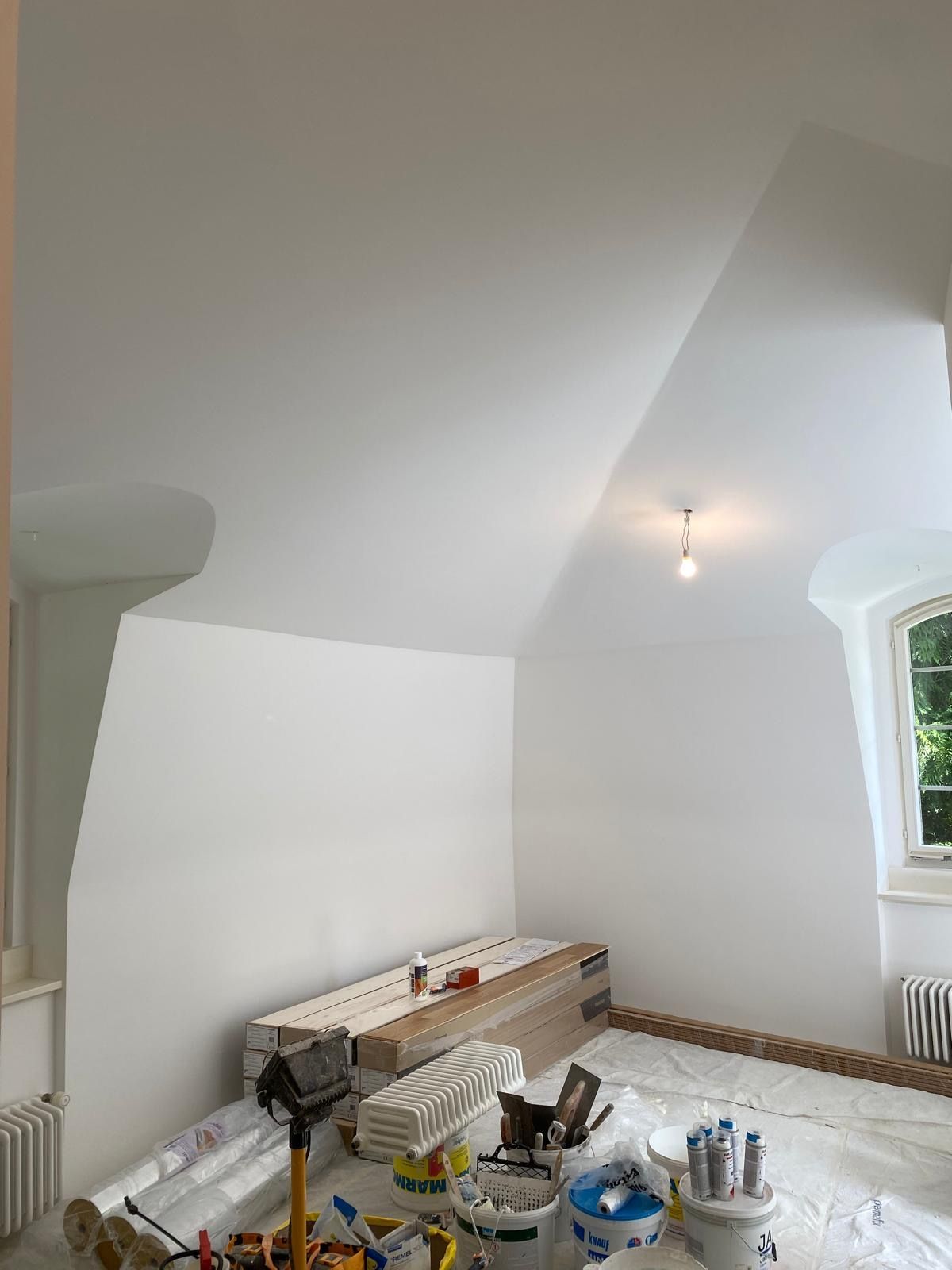 Person painting a white wall beside a paint bucket and tray, with plastic sheeting on the floor