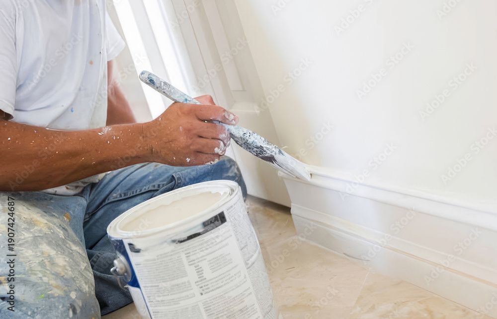 Person painting a white wall trim with a brush beside a paint bucket.
