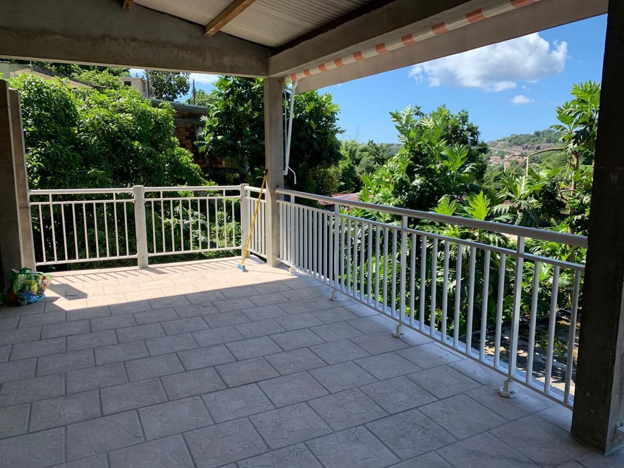 Terrasse extérieure couverte avec balustrades blanches, sol carrelé et vue sur des arbres verdoyants.