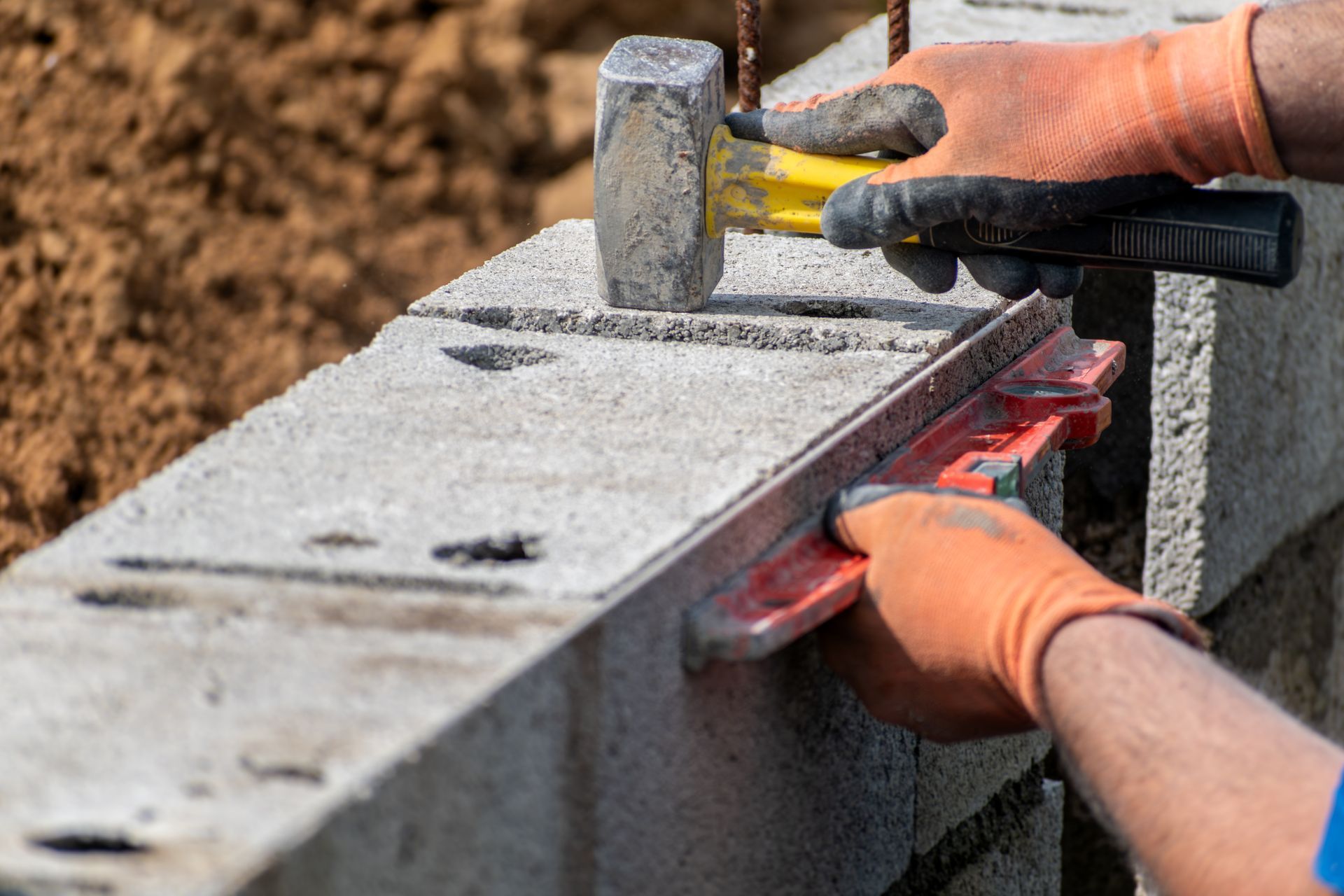 Une personne utilise un maillet pour niveler des blocs de béton avec un niveau rouge, portant des gants de travail orange.