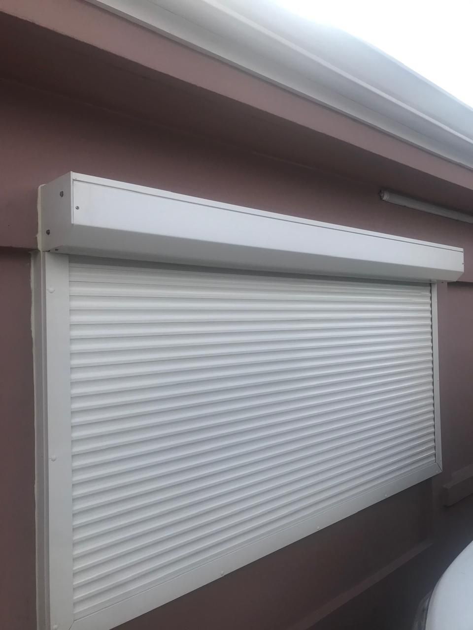 Un volet roulant extérieur blanc, monté sur un mur brun rougeâtre foncé.