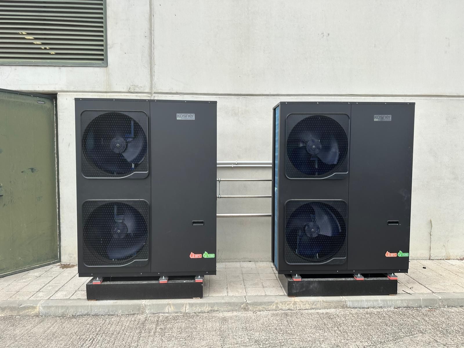 Dos bombas de calor negras, una al lado de la otra, montadas sobre plataformas negras contra una pared de hormigón.