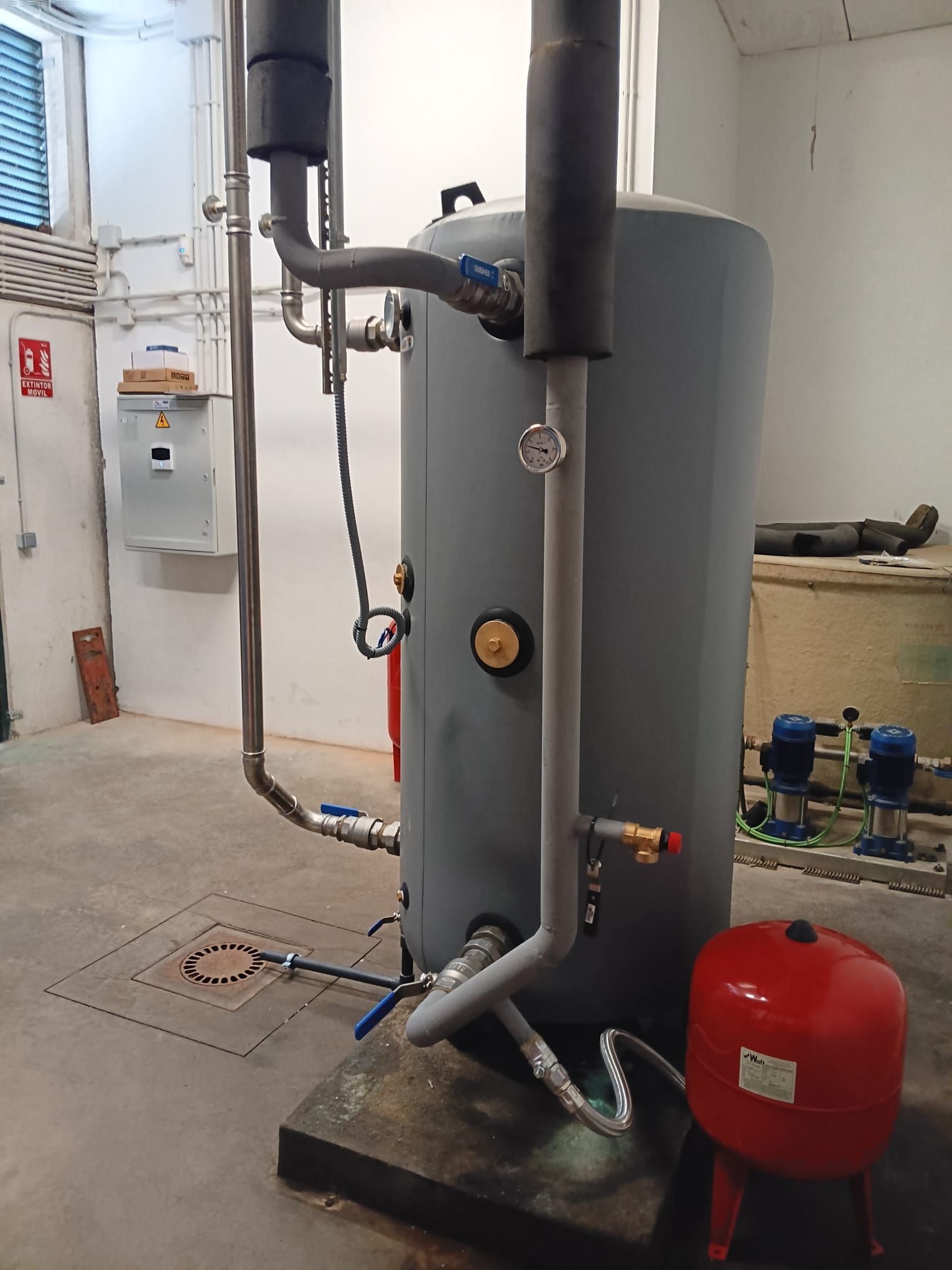 Tanque calentador de agua gris con tuberías, válvulas y un tanque de presión rojo en un cuarto de servicio.