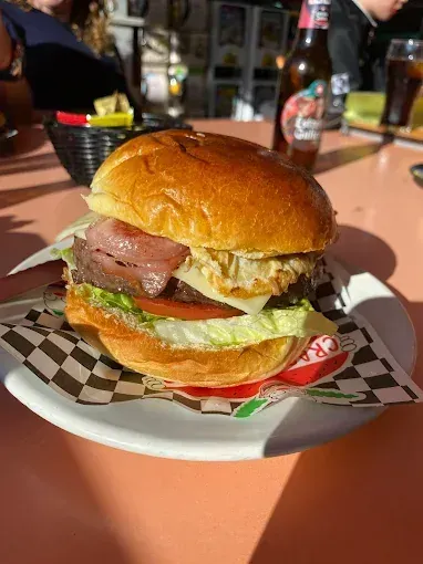 Hamburguesa en un plato con aderezos, sobre un papel a cuadros, sobre una mesa rosa. Al fondo, una botella de cerveza.