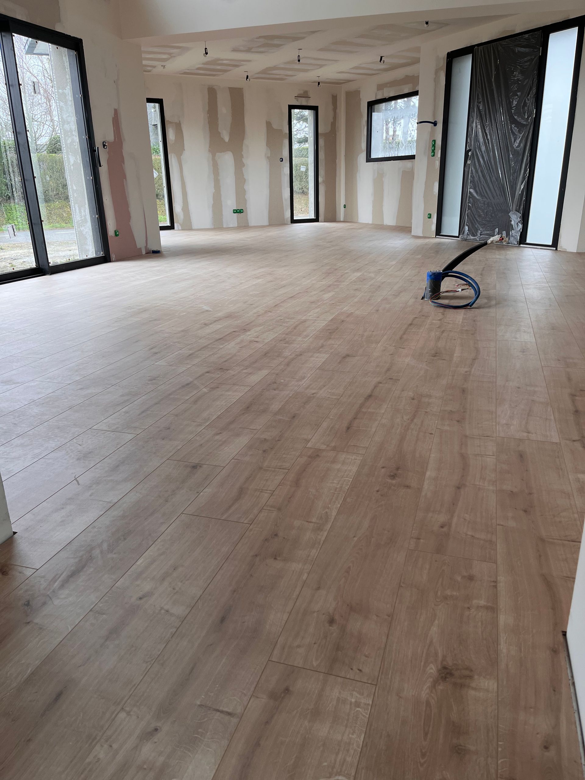 Pièce vide et inachevée, avec un parquet clair, des murs à nu, de grandes fenêtres et un outil électrique posé au sol.