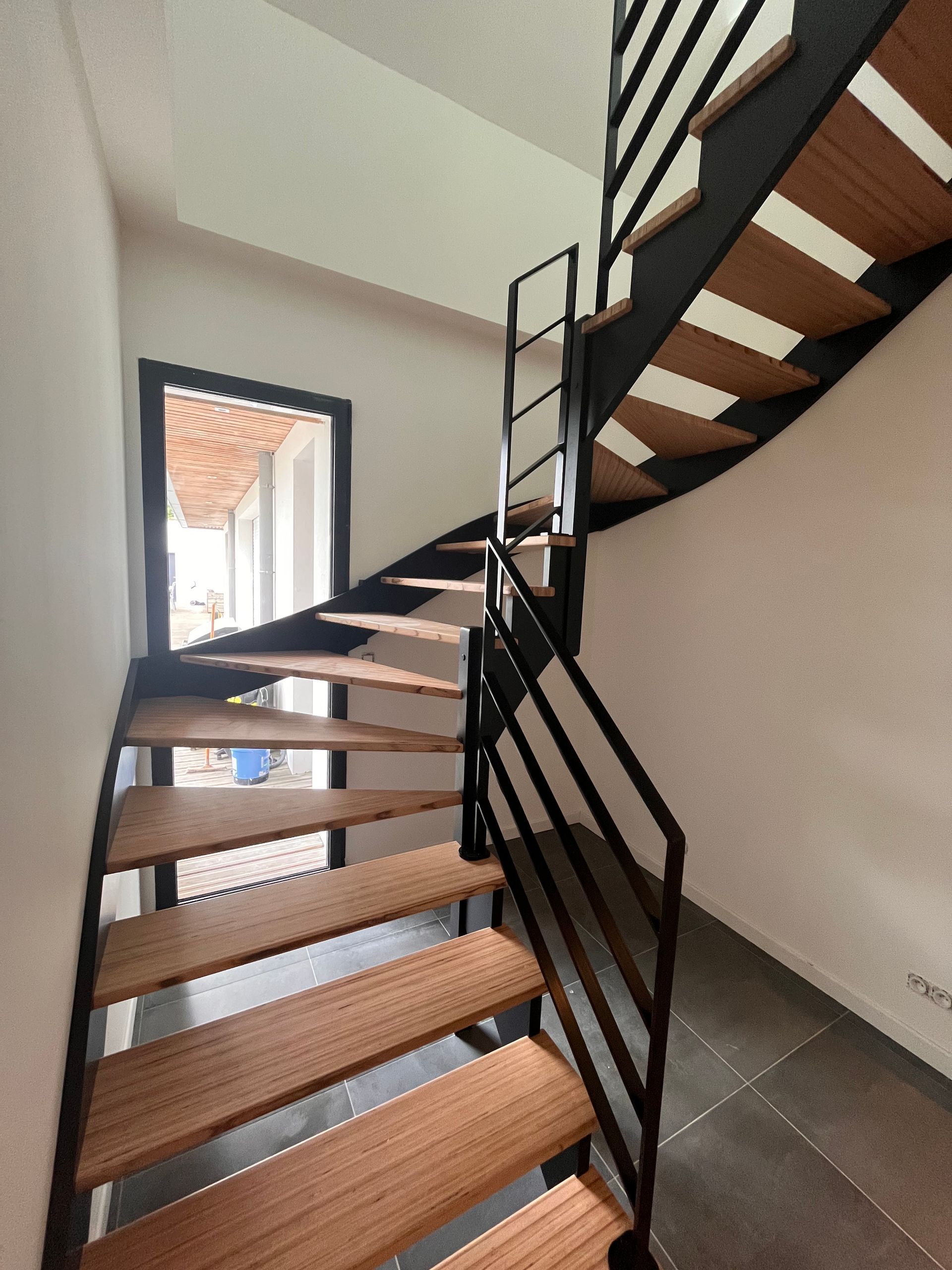 Escalier en colimaçon intérieur avec des marches en bois, une rampe en métal noir et une belle lumière naturelle.