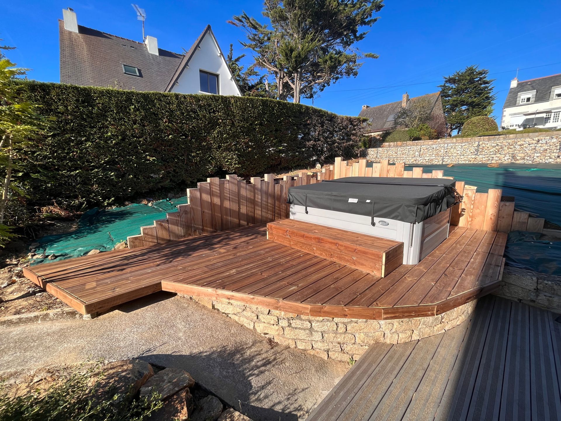Jardin avec une terrasse incurvée en briques non terminée et un bain à remous près d’une haie et de la maison