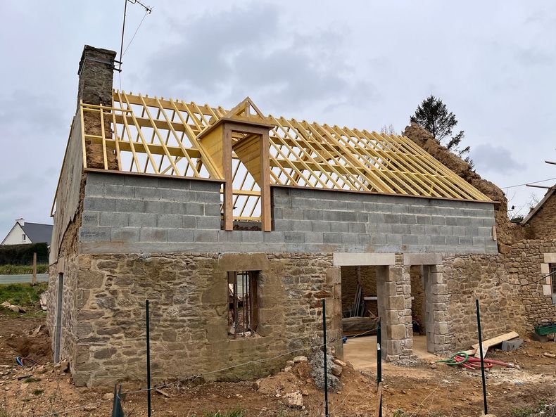 Maison en pierre en cours de construction, avec une charpente de toit jaune et des murs inachevés, sous un ciel nuageux
