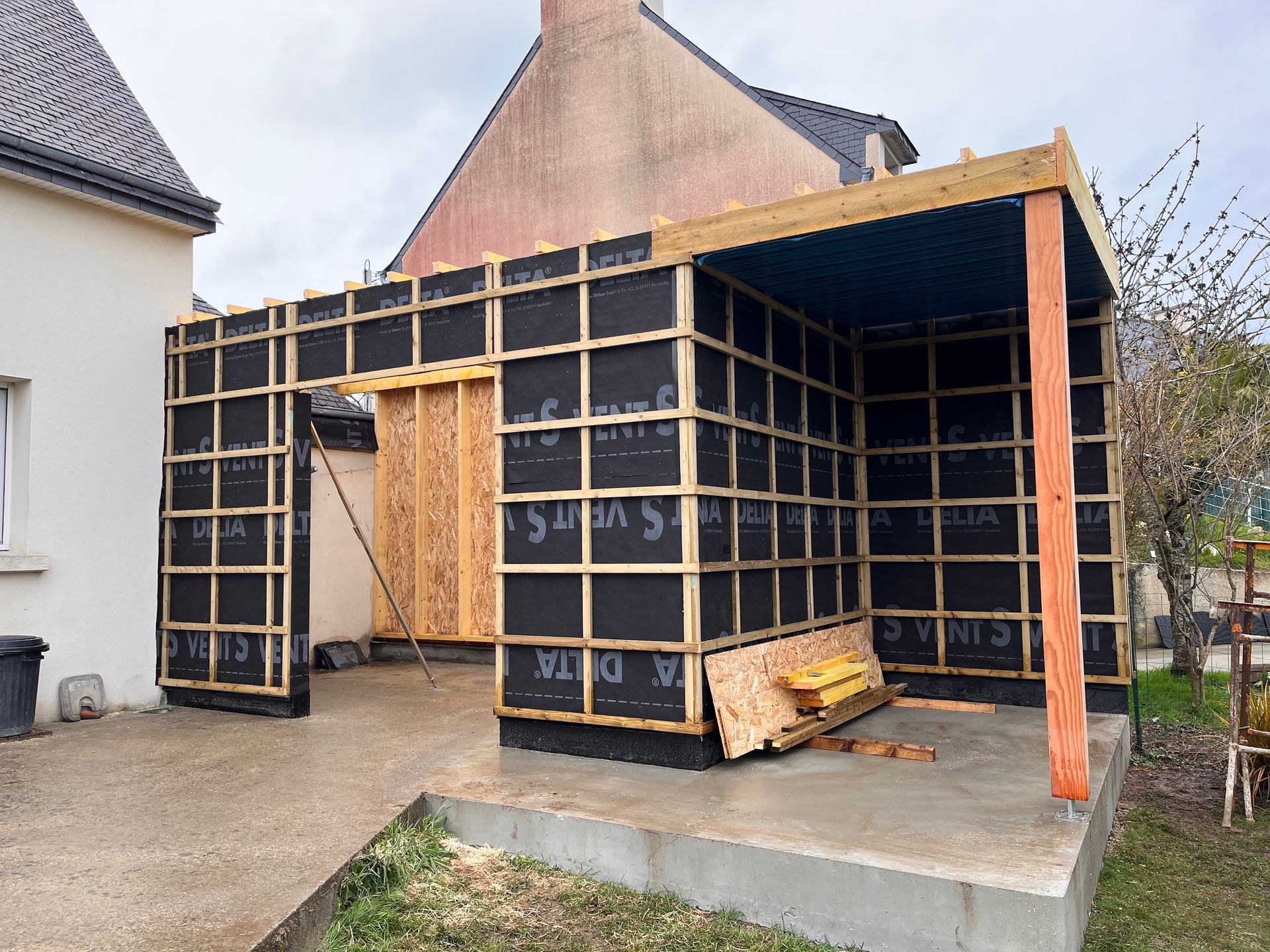 Extension de maison en construction, avec des panneaux muraux noirs et une ossature en bois, près d’une allée