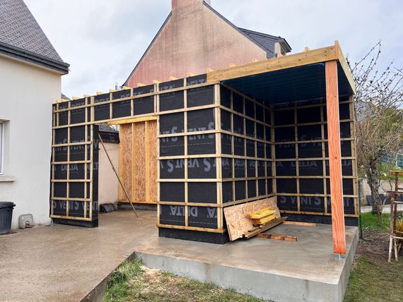 Extension à ossature en bois noir en cours de construction, accolée à une maison, avec un échafaudage et un panneau en bois posé à proximité