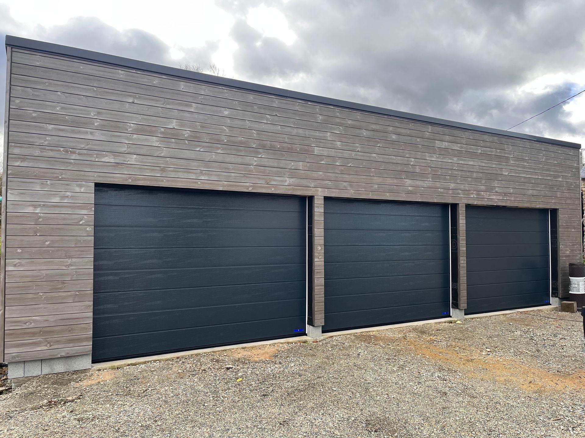 Garage moderne gris avec trois portes bleu foncé et une allée en gravier sous un ciel nuageux