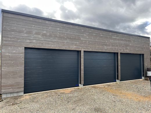 Garage moderne gris avec trois portes bleu foncé et une allée en gravier sous un ciel nuageux