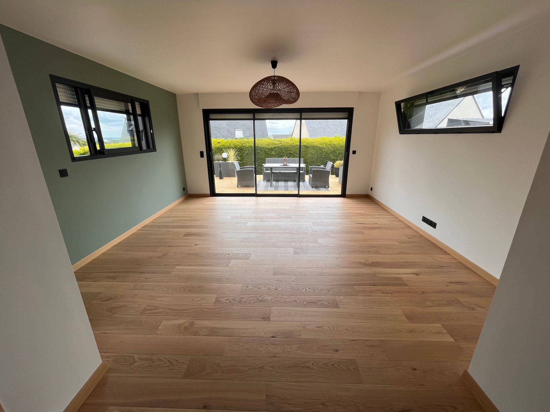 Salon moderne vide avec parquet, grandes portes vitrées et fenêtres latérales donnant sur la verdure
