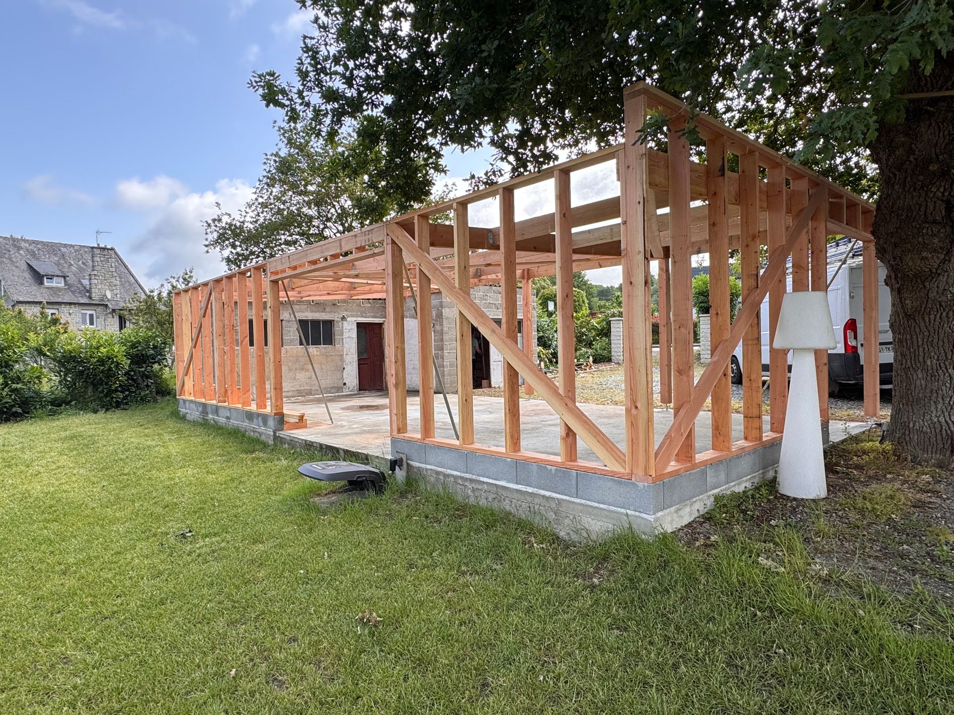 Extension à ossature en bois en construction au bord d’un jardin herbeux, avec des fondations en béton et des arbres au-dessus