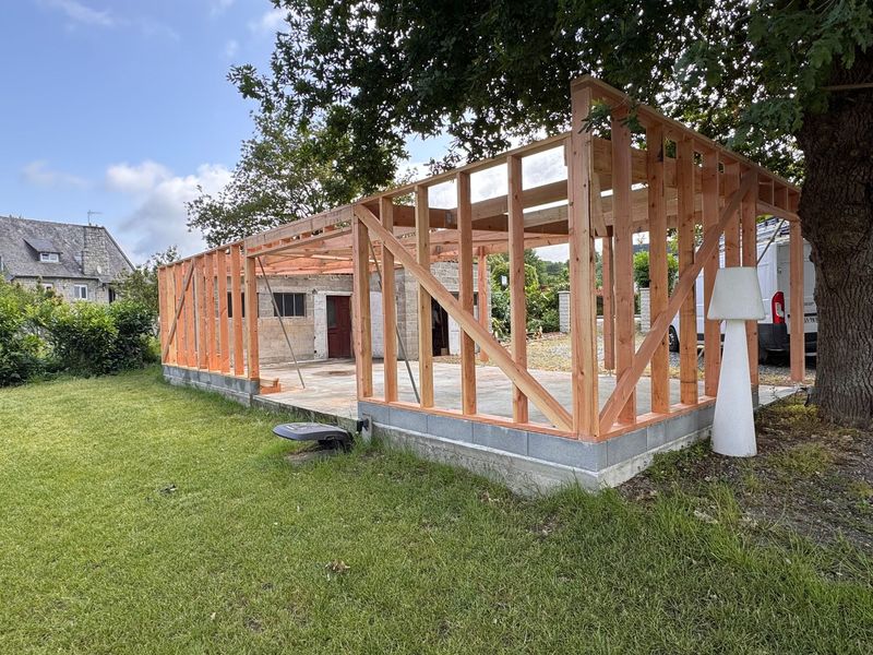 Extension à ossature bois en construction au bord d’un jardin herbeux, avec des fondations en béton et des arbres au-dessus
