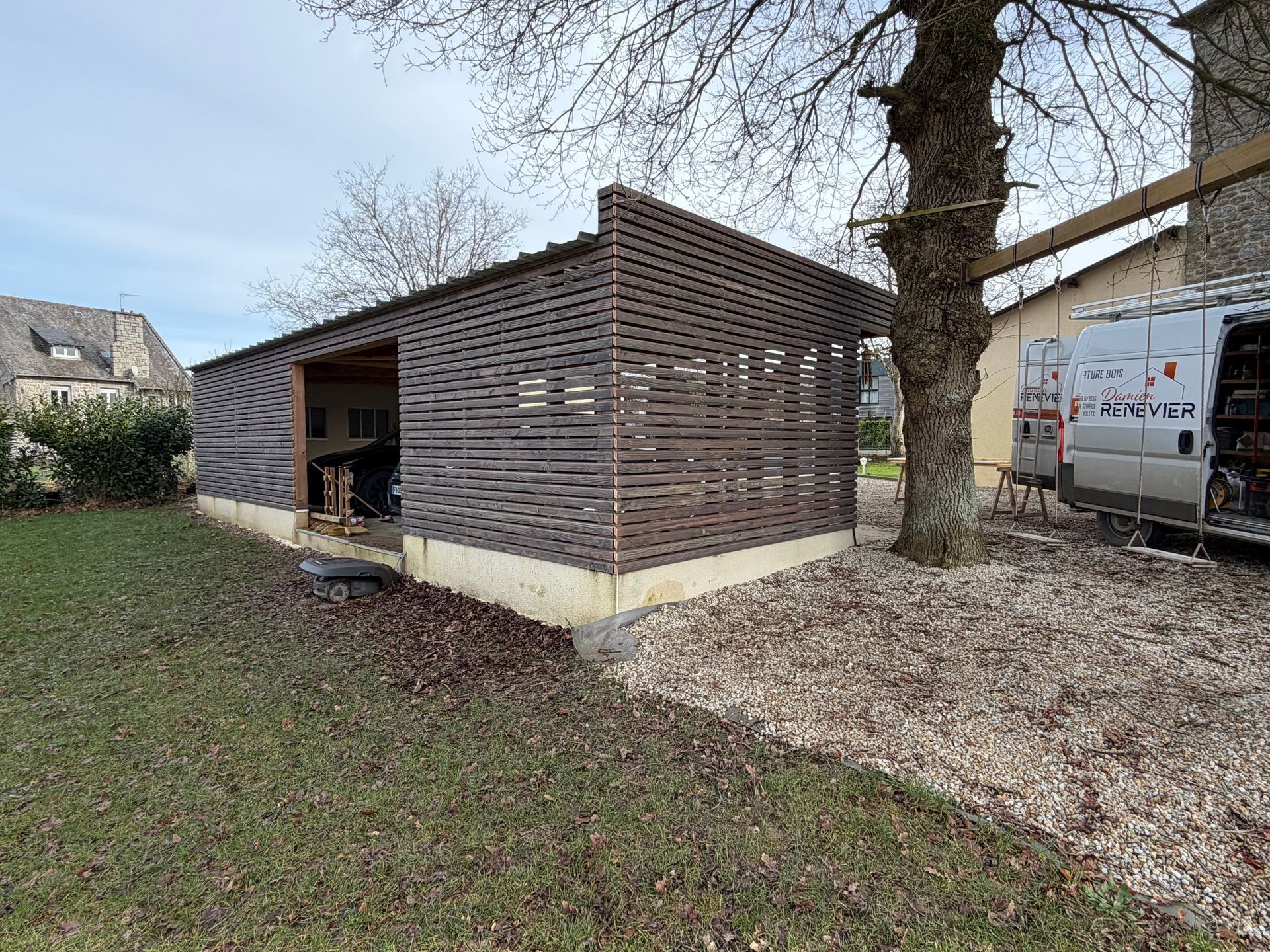 Petit abri en bois à côté d’un arbre, dans une cour gravillonnée, avec de l’herbe au premier plan et une camionnette à proximité.