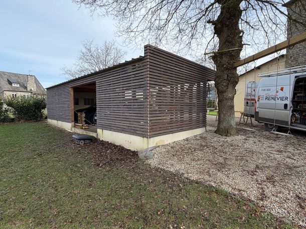Petit abri en bois à côté d’un arbre, dans une cour gravillonnée, avec de l’herbe au premier plan et une camionnette à proximité.