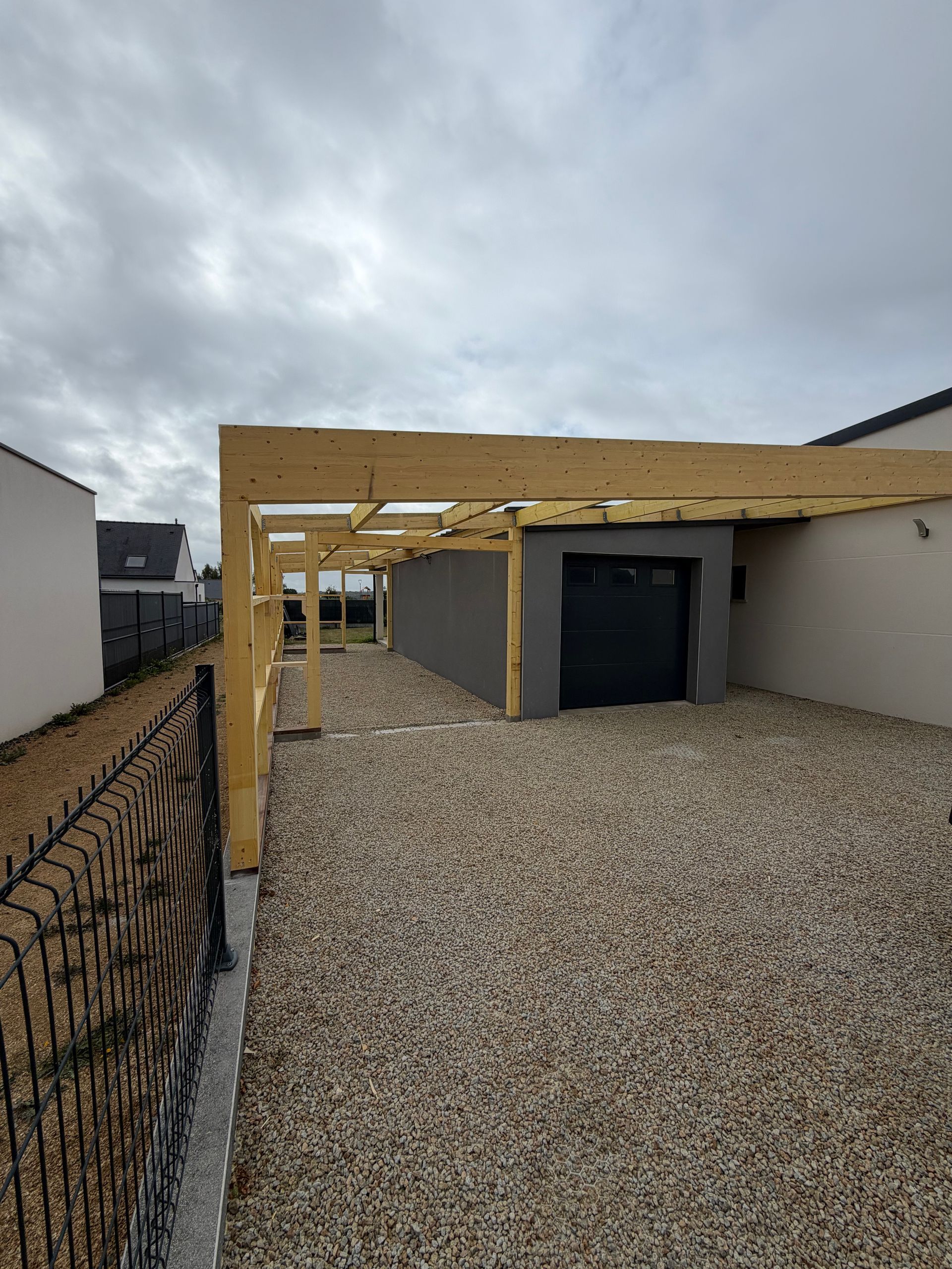Allée en gravier à côté d’une maison, avec une structure d’abri pour voiture en bois en cours de construction, par temps nuageux