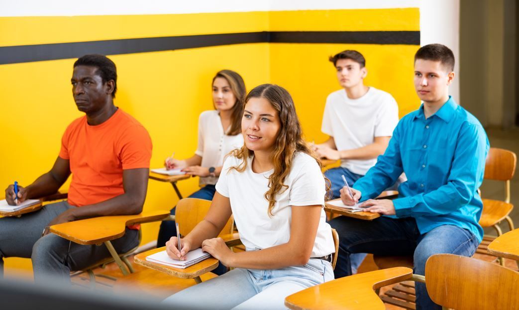 Estudiantes sentados en sus pupitres en un aula, escuchando. Pared amarilla con detalles en negro.