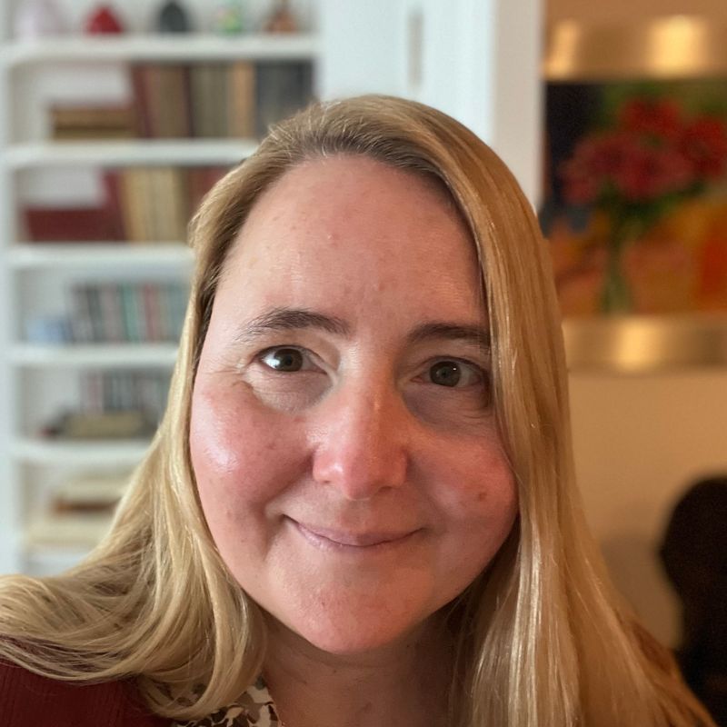 Una mujer rubia de piel clara sonríe, mirando a la cámara. Estantería y arte colorido al fondo.