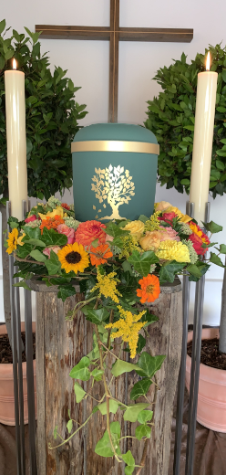 Trauerdekoration mit grüner Urne, Blumen, Kerzen und Kreuz.