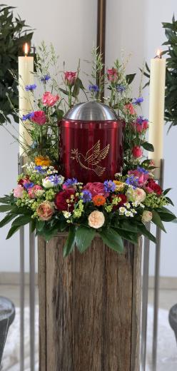Rote Urne mit Taube auf Holzsockel, umgeben von Blumen und brennenden Kerzen.