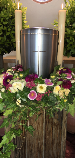 Eine silberne Urne mit einem Kreuz, umgeben von Blumen und brennenden Kerzen, auf einem Holzständer.