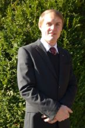 Mann im Anzug steht im Freien, die Arme verschränkt, im Hintergrund eine Hecke.