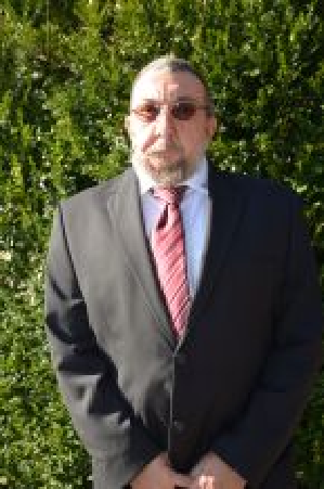 Ein Mann im Anzug mit Krawatte steht vor grünem Laub und trägt eine Brille.