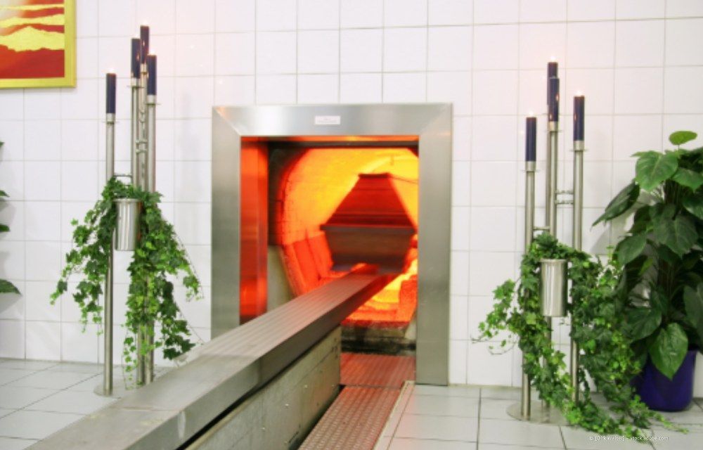 Einäscherungskammer mit orangefarben leuchtendem Innenraum, in die ein Sarg eingelassen wird. Der umgebende Bereich ist ein steriler, weiß gefliester Raum.