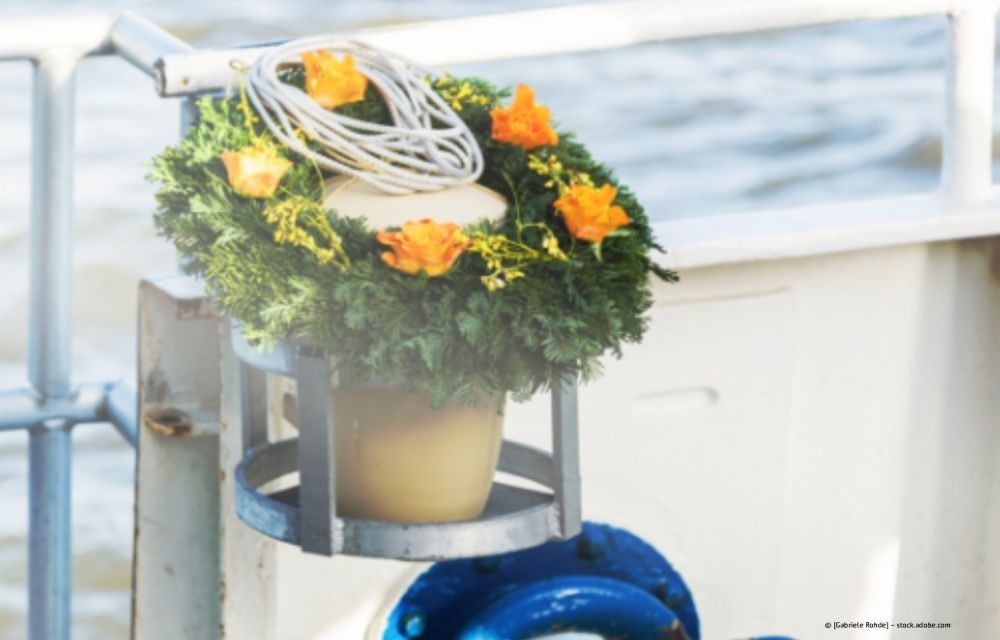 Ein Kranz mit orangefarbenen Blüten und weißem Seil auf einem Boot, vermutlich für eine Gedenkveranstaltung auf dem Wasser.