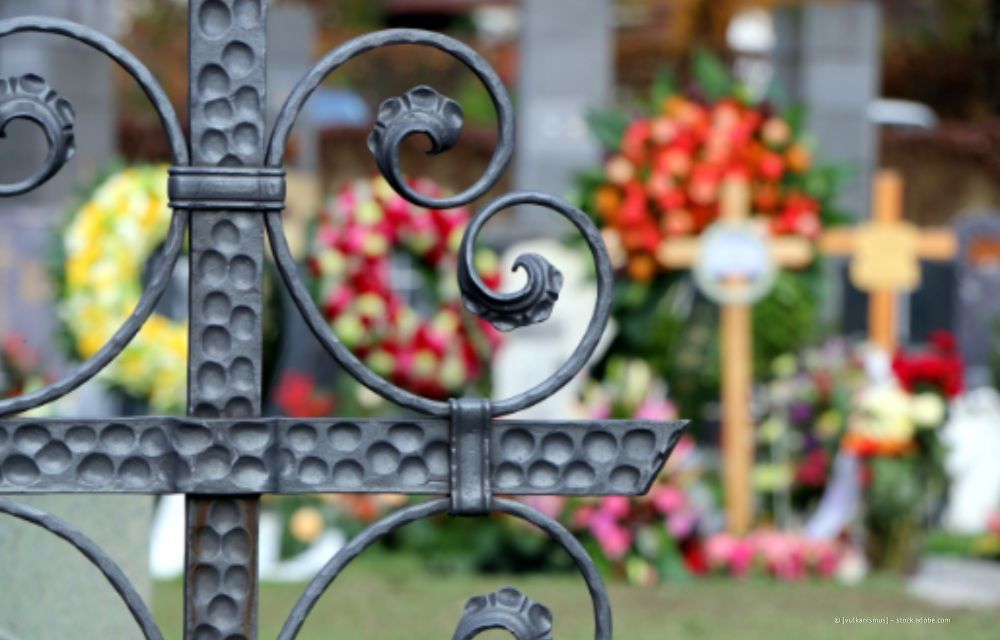 Detail eines schwarzen schmiedeeisernen Zauns vor einem Friedhof, bunte Blumenkränze und Kreuze im unscharfen Hintergrund.