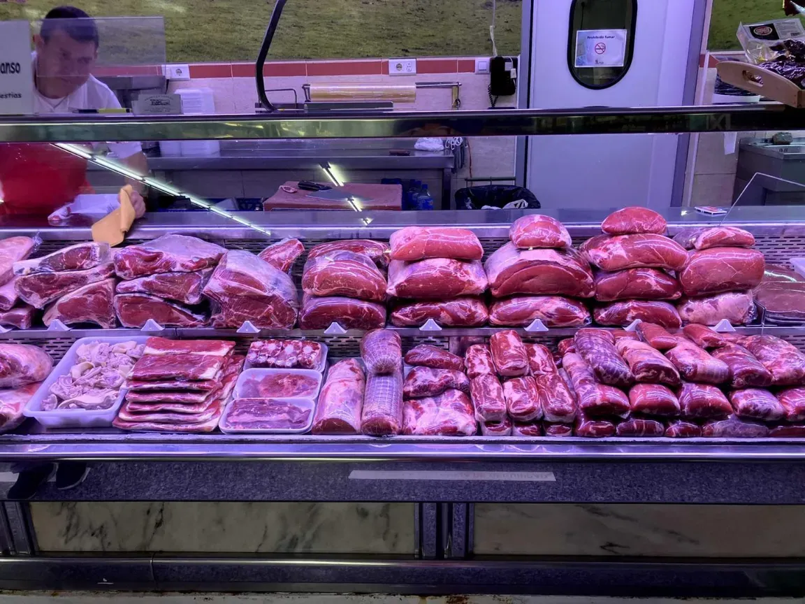 Mostrador de carne en un supermercado con bandejas de carne roja, cortes colgados en la parte superior y vitrinas iluminadas con luz rosa.
