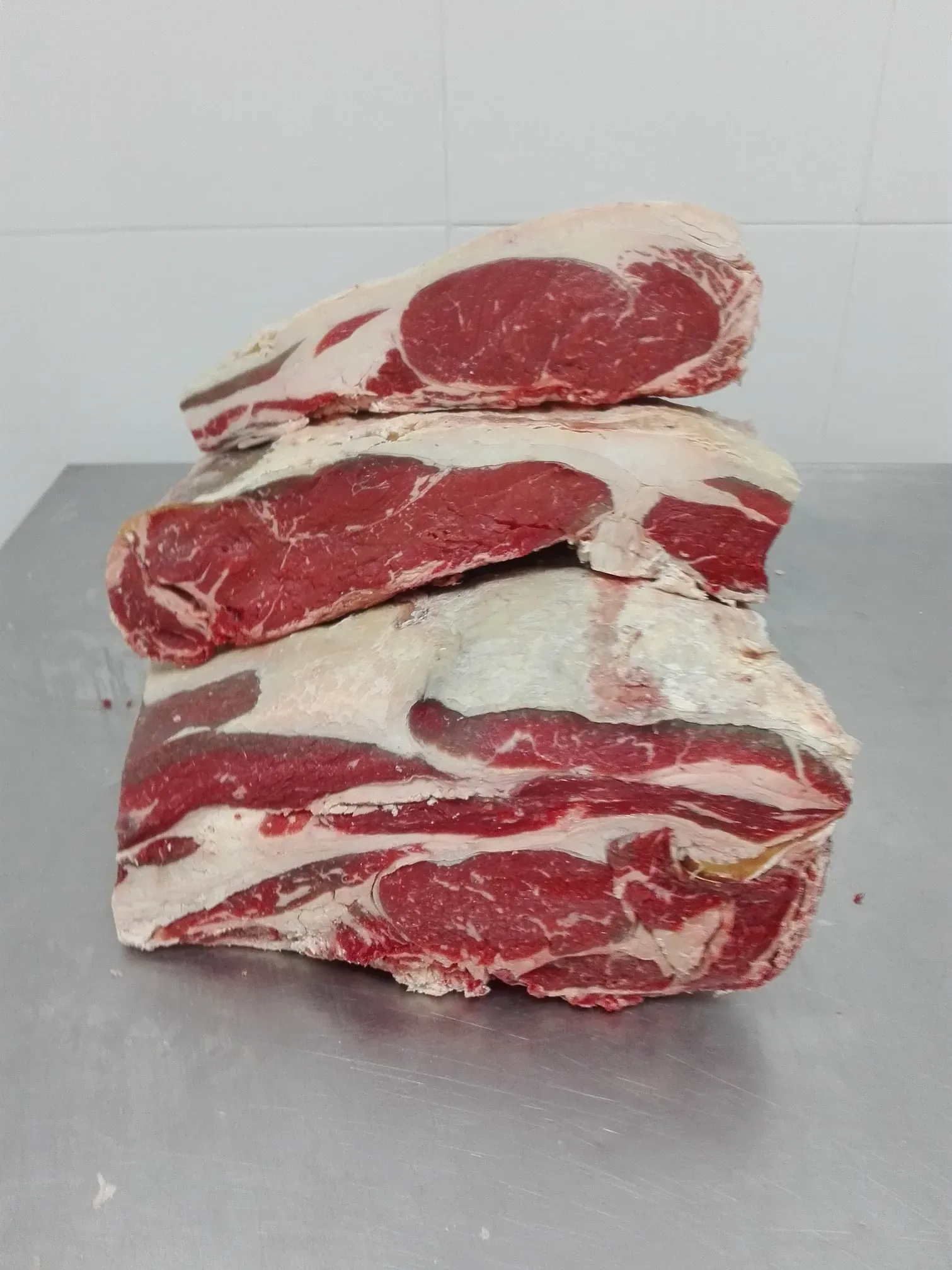 Filetes de ternera cruda apilados sobre una encimera de acero inoxidable contra una pared de azulejos blancos.