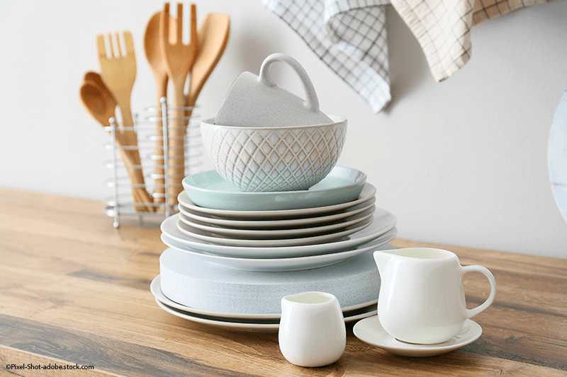 Ein Stapel weißer und hellblauer Teller mit Holzbesteck steht in einem Drahtgestell auf einer hölzernen Arbeitsplatte.