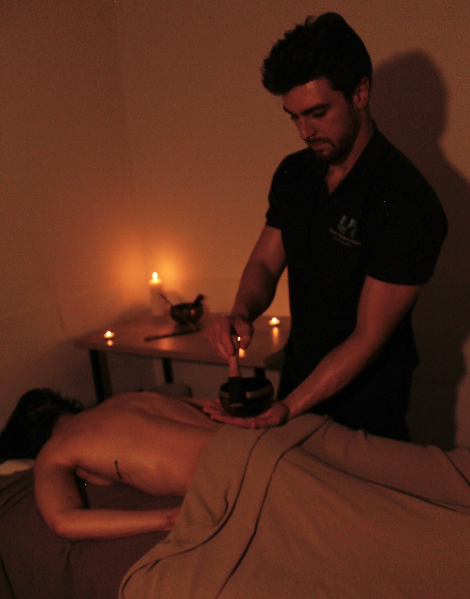 Masajista dando un masaje con piedras calientes a un cliente en una sala con poca luz y velas