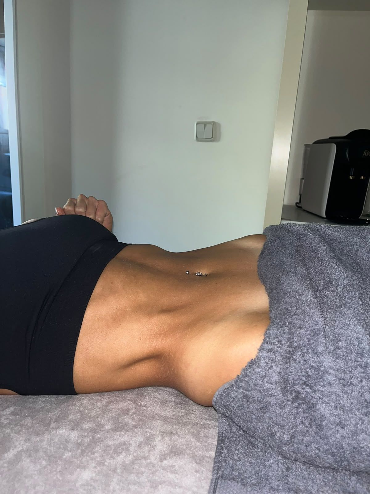 Una persona tumbada en una cama, con la espalda y la cintura al descubierto y una manta gris cerca.