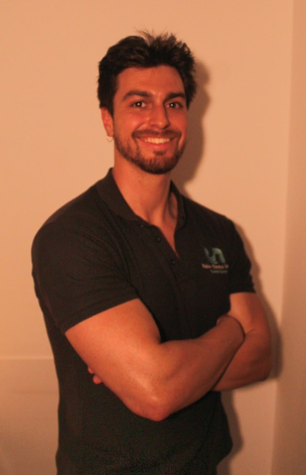 Hombre sonriente con los brazos cruzados, vestido con una camisa oscura y de pie frente a una pared beige.