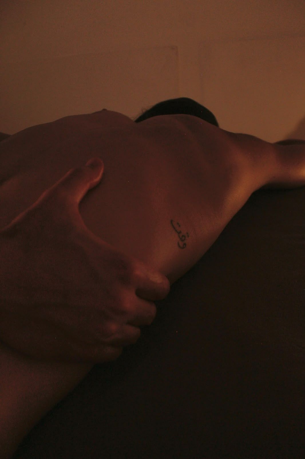Una mano apoyada sobre un hombro descubierto, con una iluminación cálida y tenue y un pequeño tatuaje a la vista