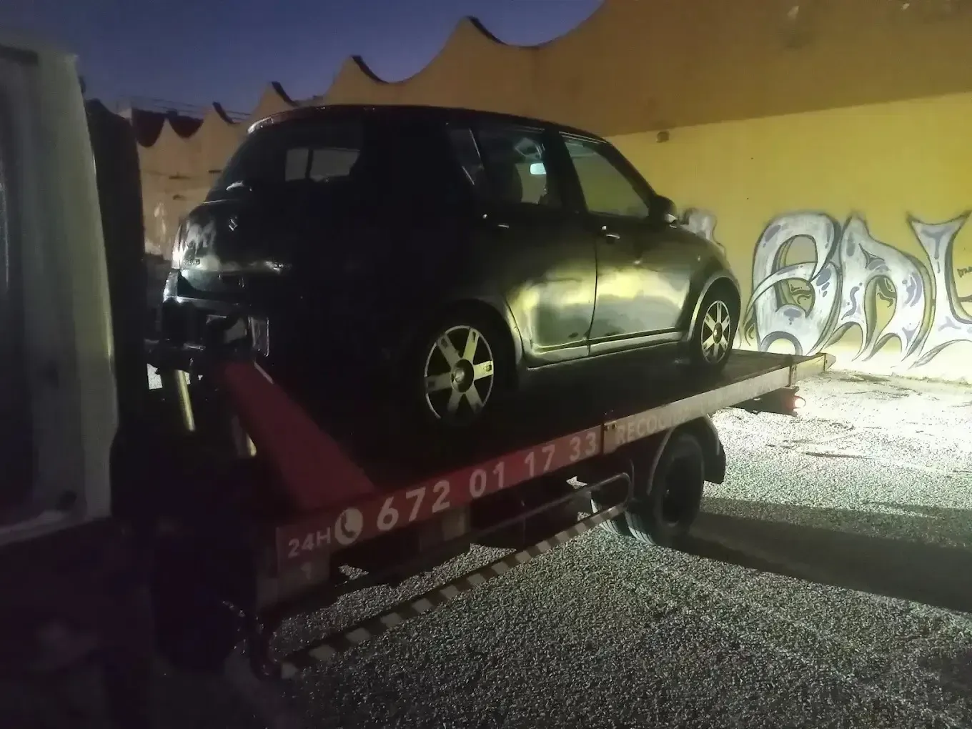 Un coche compacto negro subido a la plataforma de una grúa frente a un muro con grafitis.