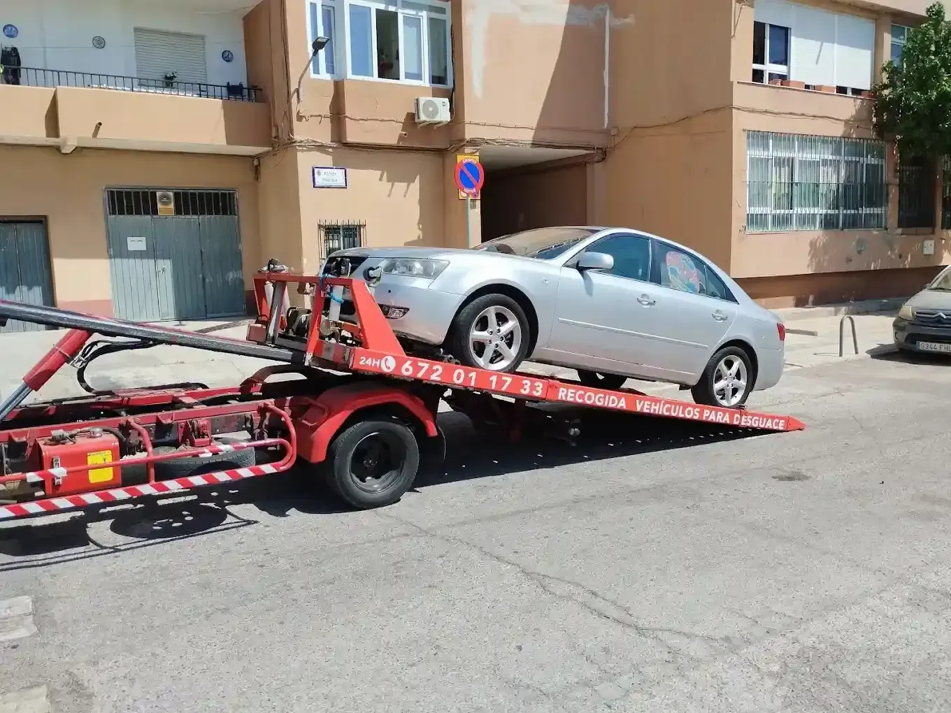 Un sedán plateado está siendo cargado en una grúa de plataforma estacionada en una calle residencial.