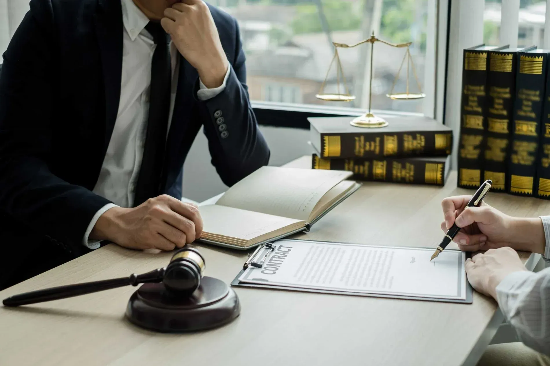 Un abogado de traje consulta con un cliente en un escritorio, revisando documentos y firmando. Se ven libros legales, mazo y balanza de justicia.